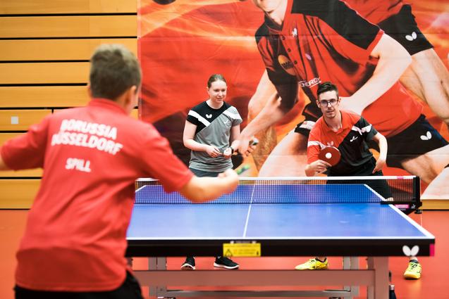 Drei Tischtennisspieler stehen an einem Tisch mit einer blauen Platte, bereit für ein Spiel in einer Sporthalle.