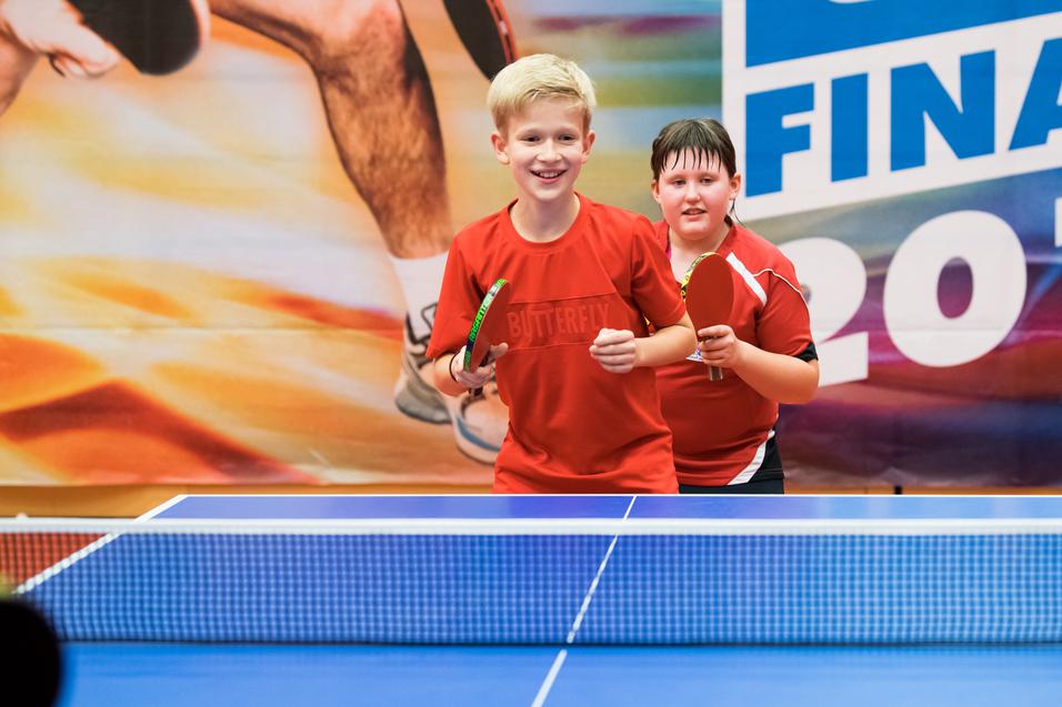 Zwei Kinder in roten Trikots stehen lächelnd an einem Tischtennistisch mit Schlägern in den Händen.