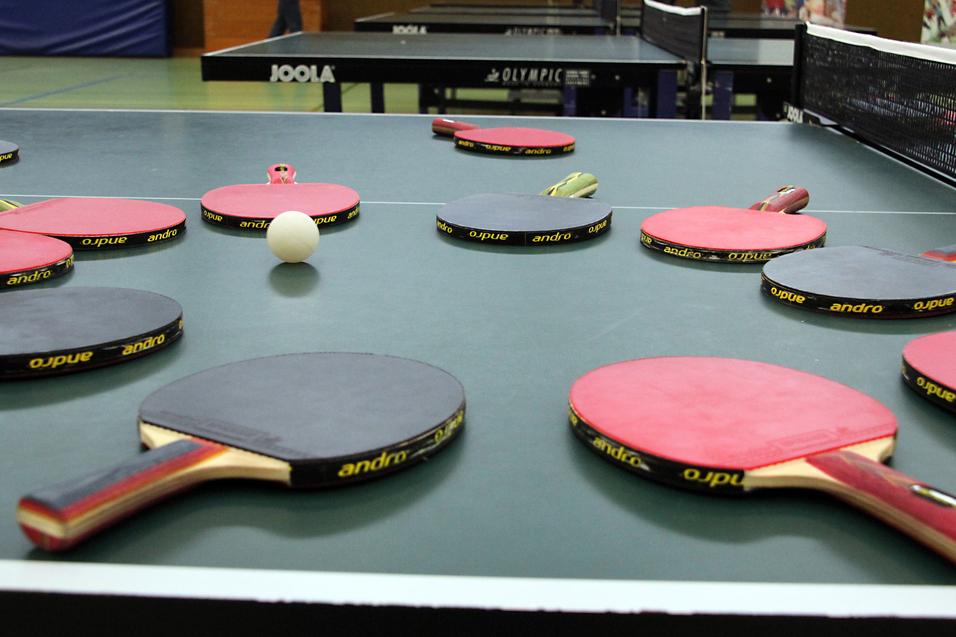 Mehrere Tischtennisschläger in verschiedenen Farben und ein Tischtennisball liegen auf einer grünen Tischtennisplatte.