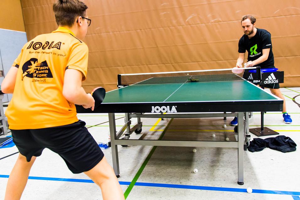 Zwei Tischtennisspieler trainieren an einem Tischtennistisch in einer Sporthalle, mehrere Bälle liegen auf dem Boden.