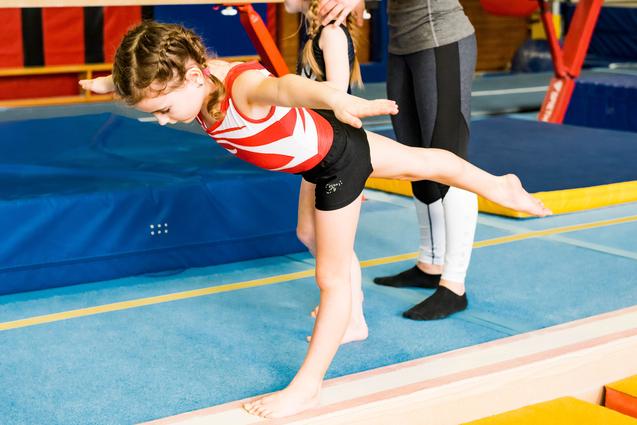 Zwei Mädchen trainieren in einer Turnhalle; eines balanciert auf einem Schwebebalken, während das andere unterstützt.