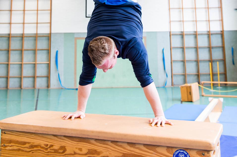 Sportler führt einen Handstand auf einer Kiste in einer Turnhalle durch, im Hintergrund sind Turngeräte sichtbar.
