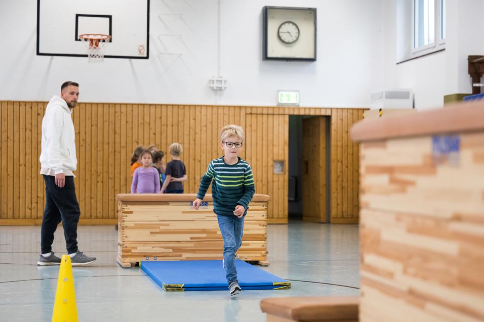 Ein Junge in gestreiftem Pullover läuft über eine Übungsfläche, während Erwachsene und Kinder im Hintergrund aktiv sind.