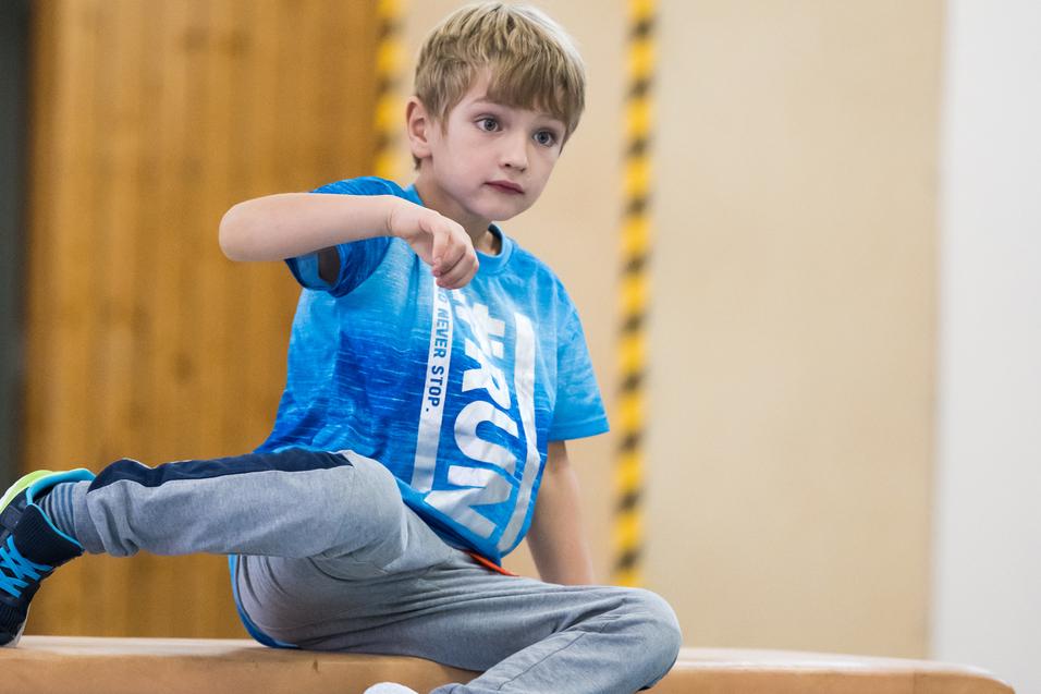 Junge sitzt auf einem Turngerät, trägt ein blaues T-Shirt und graue Sporthose, während er nachdenklich schaut.