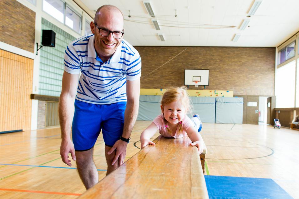 Mann lehnt am Schwebebalken, während ein lächelndes Mädchen darauf liegt, in einer hellen Sporthalle.