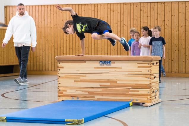 Ein Junge springt über ein Turngerät, während eine Gruppe Kinder und ein Trainer zuschauen. Sporthalle mit Holzverkleidung.