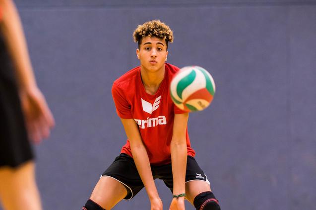 Junger Volleyballspieler in rotem Shirt bereit, einen bunten Volleyball zu empfangen, auf einem Holzsportboden.