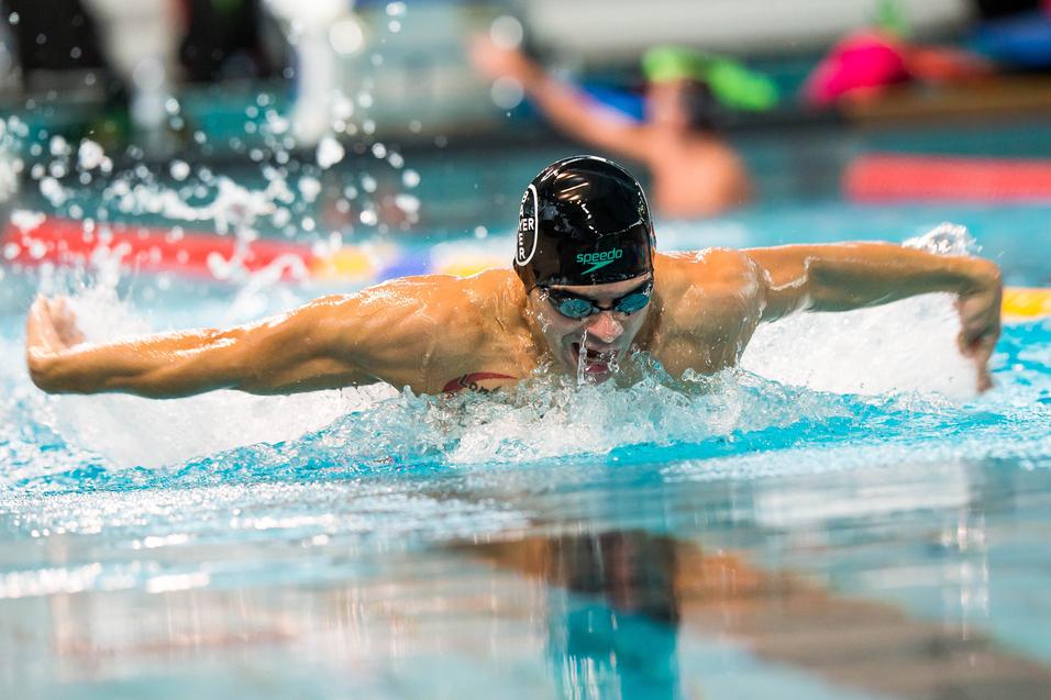 Schwimmer im Wettkampf bei der Schmetterlingsbewegung in einem Hallenbad, Wasser spritzt um ihn herum.