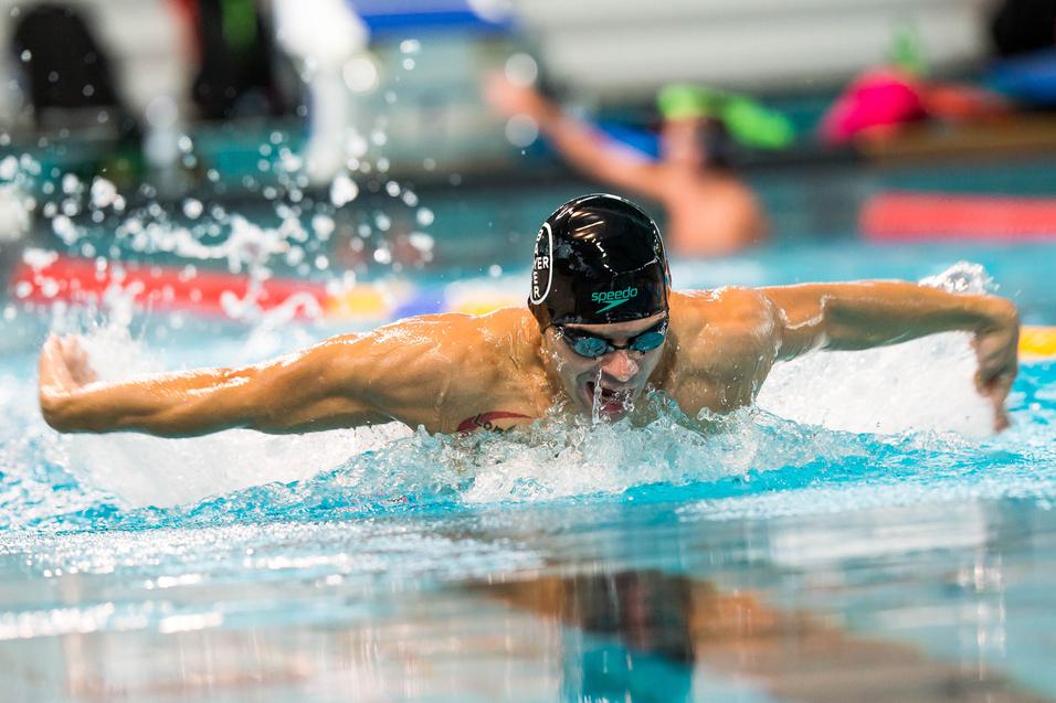 Schwimmer im Wettkampf bei der Schmetterlingsbewegung in einem Hallenbad, Wasser spritzt um ihn herum.