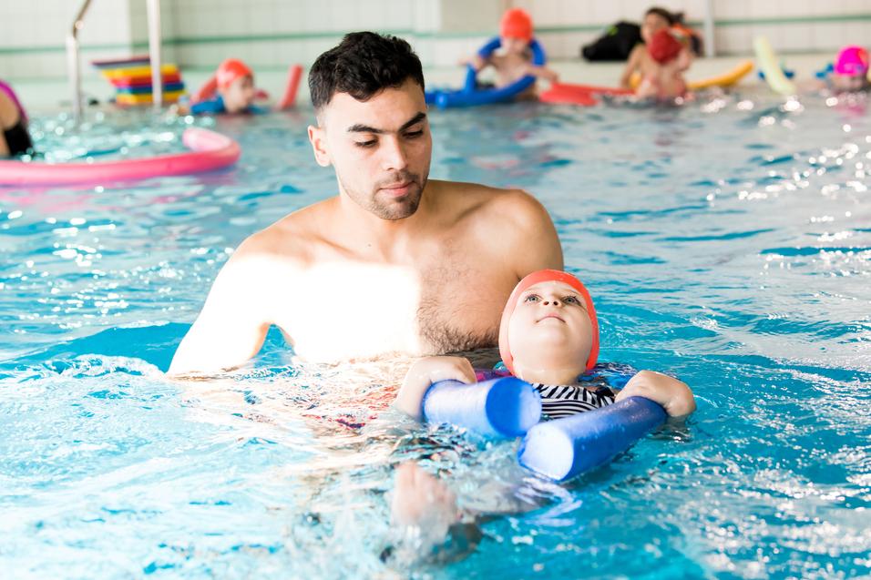 Mann unterstützt ein Kind mit Schwimmhilfen im Wasser eines Schwimmbads, während andere Schwimmer im Hintergrund sind.