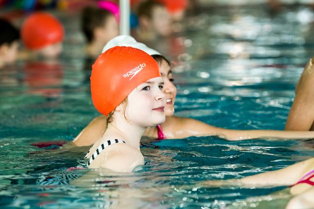 Mädchen mit oranger Schwimmhaube schwimmt in einem Hallenbad, im Hintergrund weitere Kinder im Wasser.