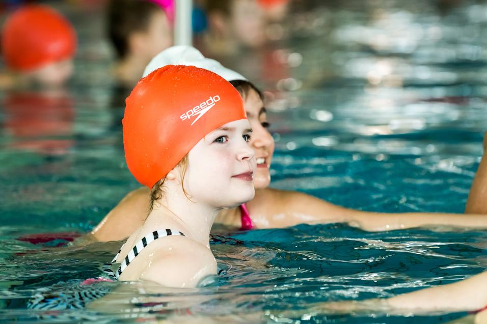 Mädchen mit oranger Schwimmhaube schwimmt in einem Hallenbad, im Hintergrund weitere Kinder im Wasser.
